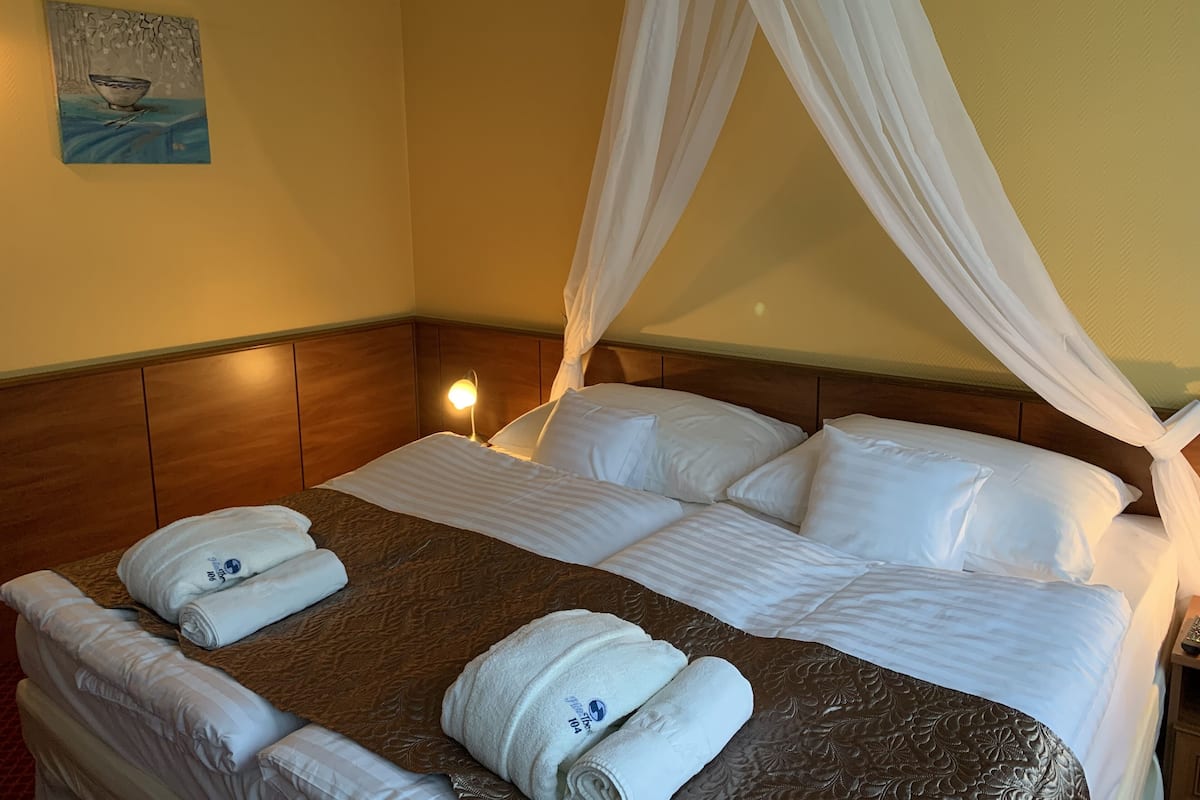 Ein ordentlich bezogenes Bett mit weißen Bettwäsche und einer braunen, gemusterten Tagesdecke, ein Nachttisch mit Lampe und ein Gemälde an der Wand.