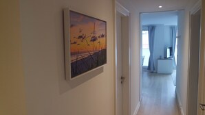 A hallway with a framed painting of a sunset, leading to a room with a bed and a window. - Strandperle (Juist)
