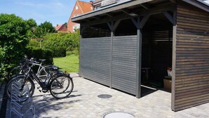 A covered bike parking area with two bicycles parked, a building with a sloped roof, and a clear sky. - Strandperle (Juist)