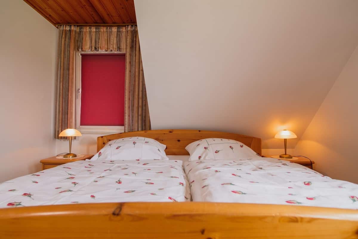 A double bed with floral bedding, two bedside lamps, a wooden headboard, a window with curtains, and a red wall.