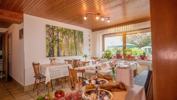 A dining area with wooden ceiling, white walls, and a large window with plants. Tables are set with white tablecloths and various dishes. - Bauernhof Pension Wiebelhaus Mester (Lennestadt)