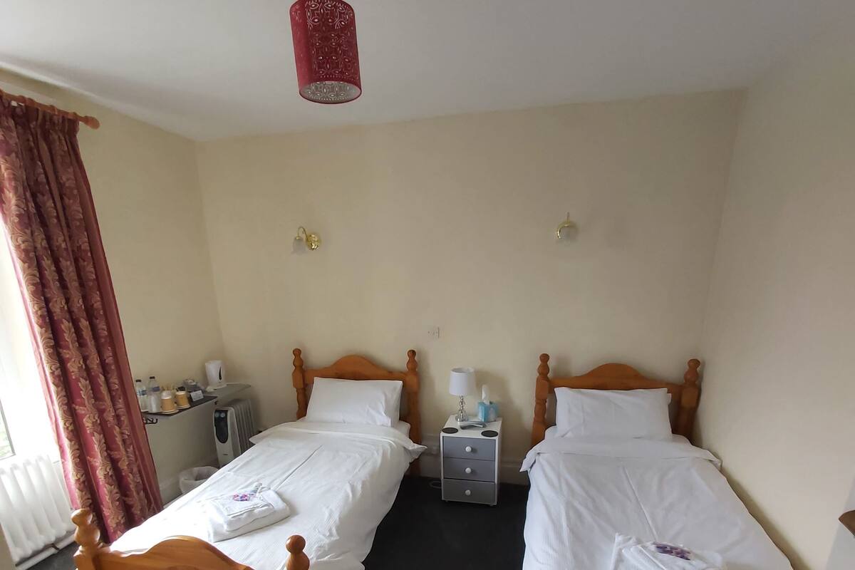 Une chambre d’hôtel avec deux lits en bois, une petite table de chevet avec une lampe, un radiateur et un luminaire rouge suspendu au plafond.