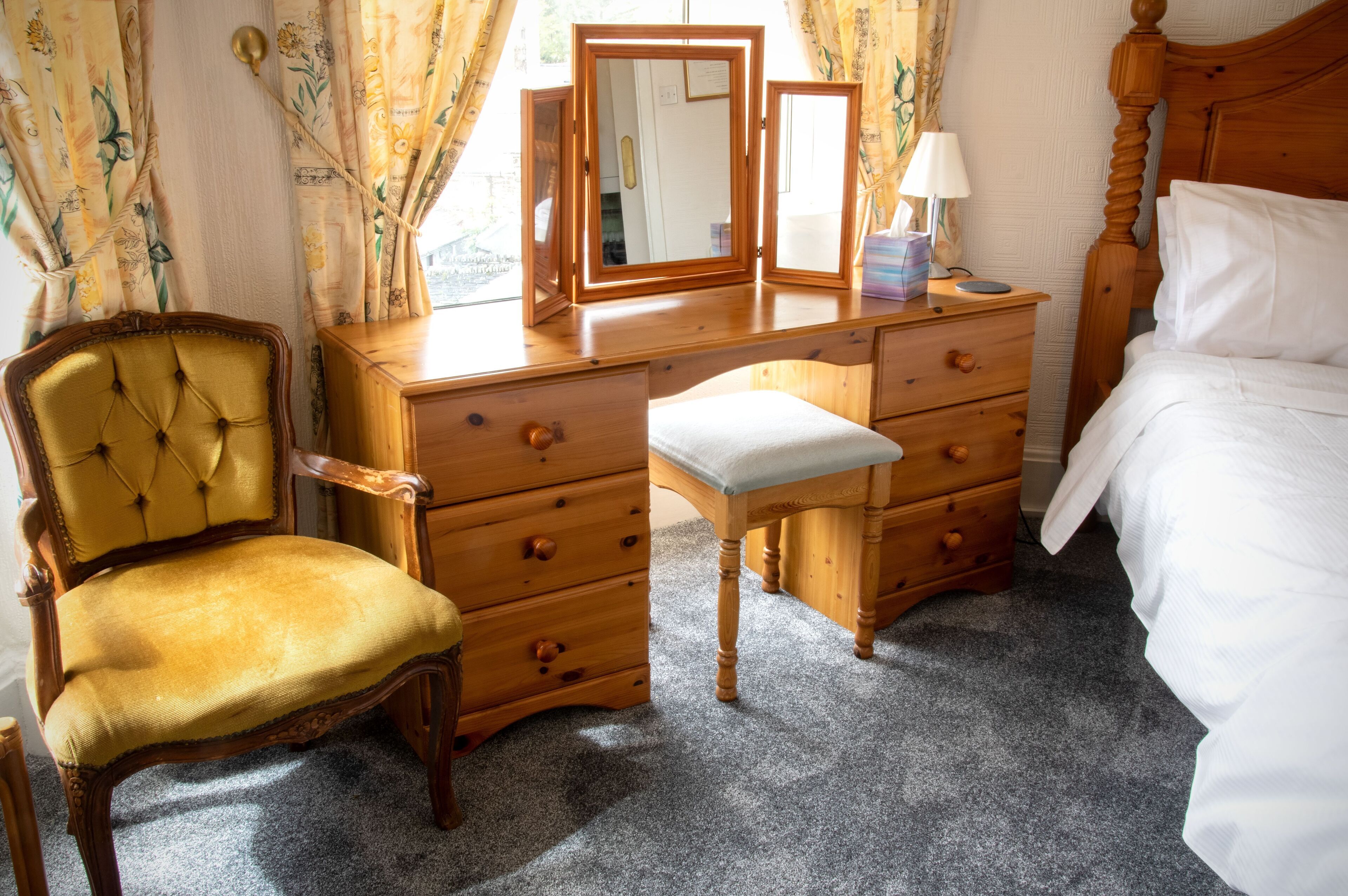 A bedroom with a wooden dresser, a mirror, a bedside table with a lamp, a chair, and a bed.