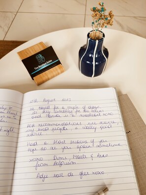 A notebook with handwritten notes, a business card, and a vase with dried flowers on a white surface. - Tins Athens (Athens)
