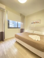 Ένα υπνοδωμάτιο με ένα μεγάλο κρεβάτι, ένα παράθυρο με κουρτίνες, ένα κλιματιστικό τοποθετημένο στον τοίχο και έναν πίνακα που απεικονίζει την πανοραμική θέα της πόλης.