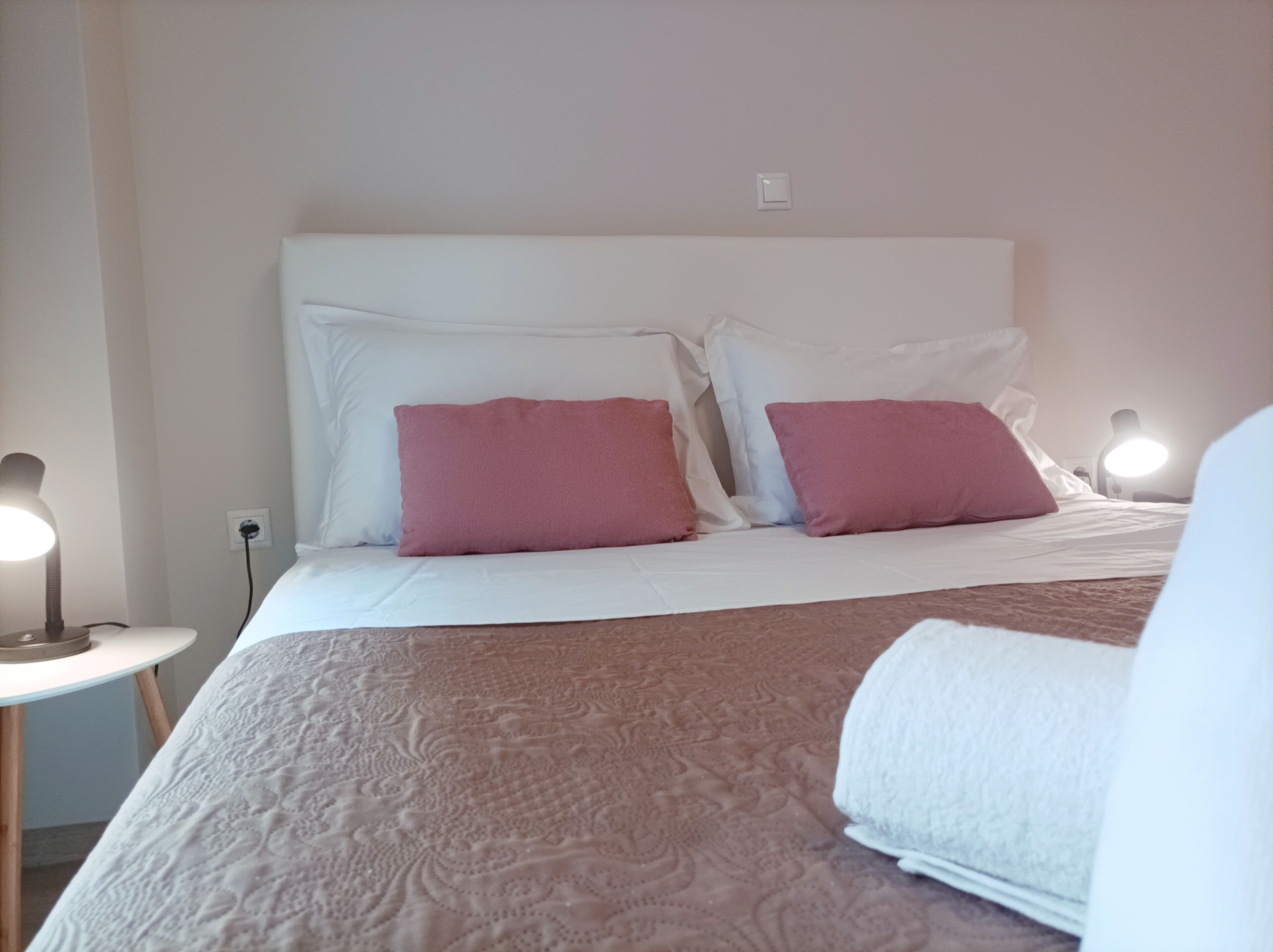 A neatly made bed with white and pink pillows, a bedside lamp, and a power outlet on the wall.