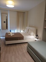 Quarto de hotel moderno com uma cama grande, mesa de cabeceira com abajur, uma mesinha e uma janela com cortinas.