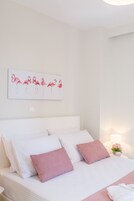 Uma cama bem arrumada com roupa de cama branca e rosa, uma obra de arte na parede com flamingos e uma pequena mesa de cabeceira com uma luminária e flores.