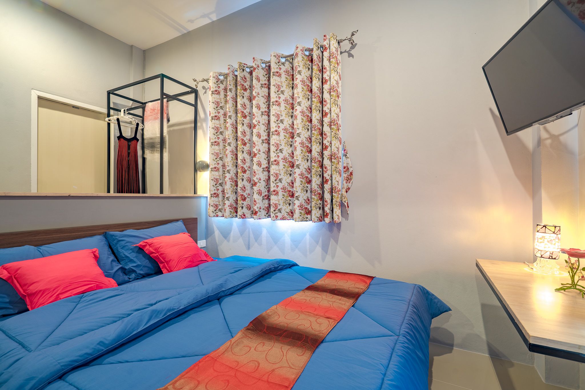 Une chambre à coucher avec un lit, une télévision, une table de chevet, un miroir et une fenêtre ornée de rideaux à motifs floraux.
