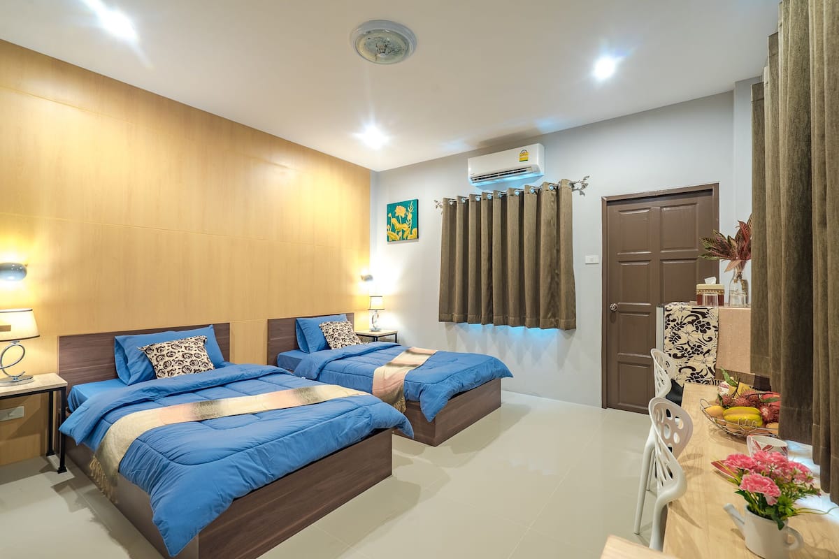 A hotel room with two beds, a dining table with chairs, a decorative vase, and an air conditioning unit.