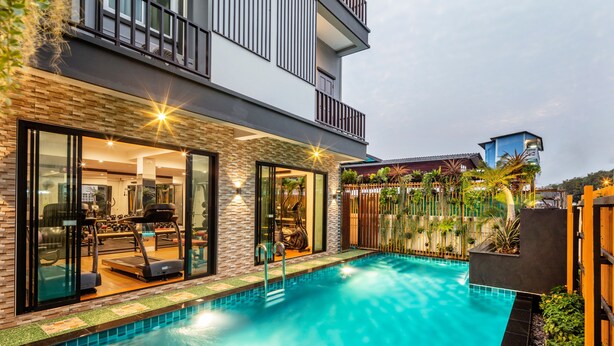 A modern pool area with a glass-enclosed gym, a two-story building with balconies, and a well-maintained garden. - Penyos Residence (Bangkok)