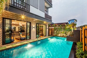 Un área de piscina moderna con un gimnasio de cristal, un edificio de dos pisos con balcones y un jardín bien cuidado.