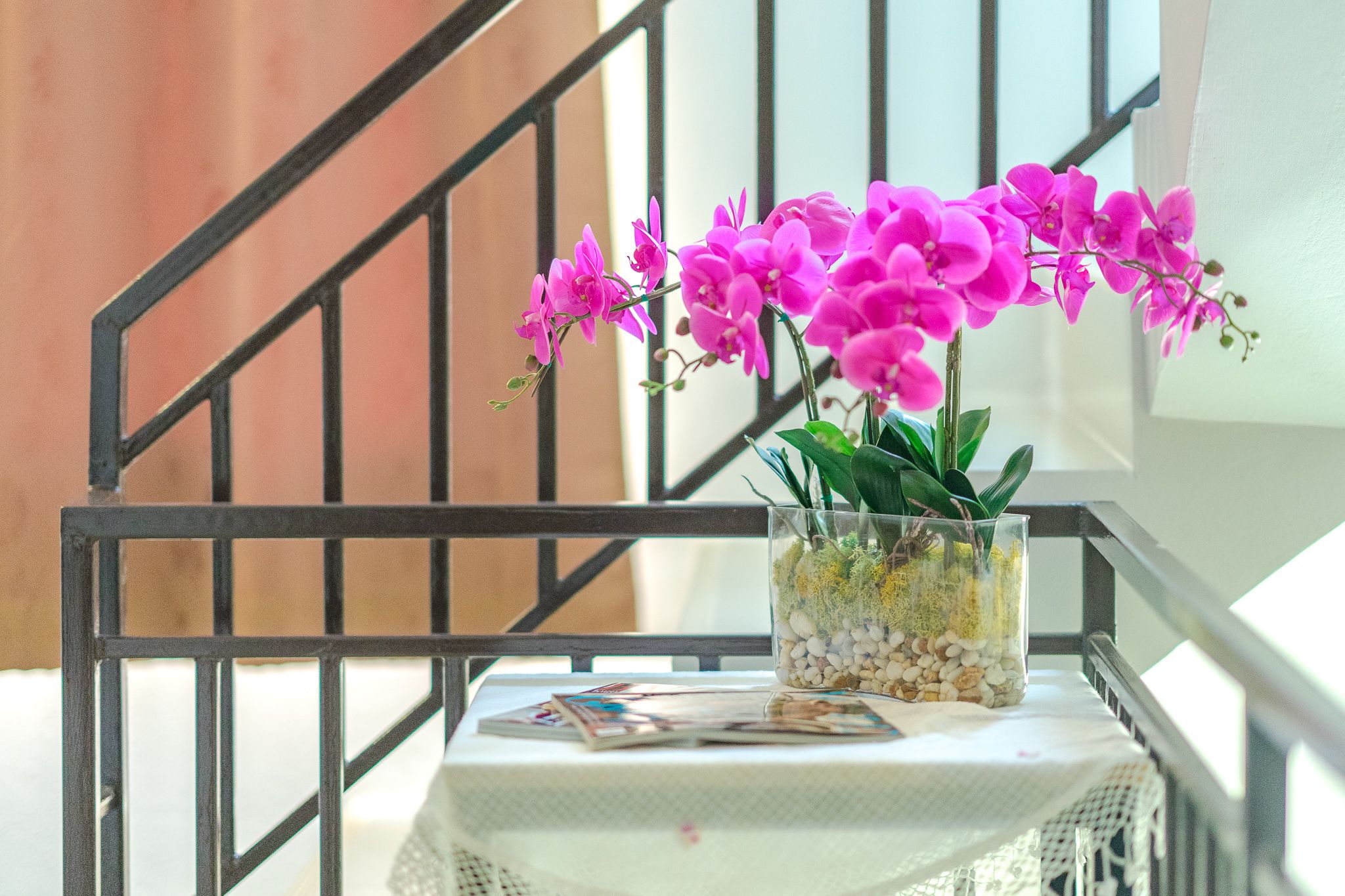 Un florero de vidrio con orquídeas rosadas sobre una mesa con revistas, frente a una barandilla de escalera.