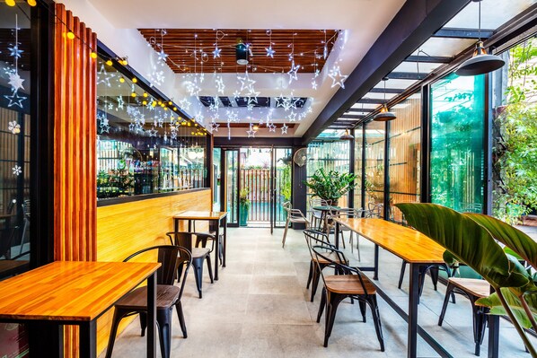 Un café moderno con mesas y sillas de madera, lámparas colgantes en forma de estrella y grandes ventanales con vistas a zonas verdes.
