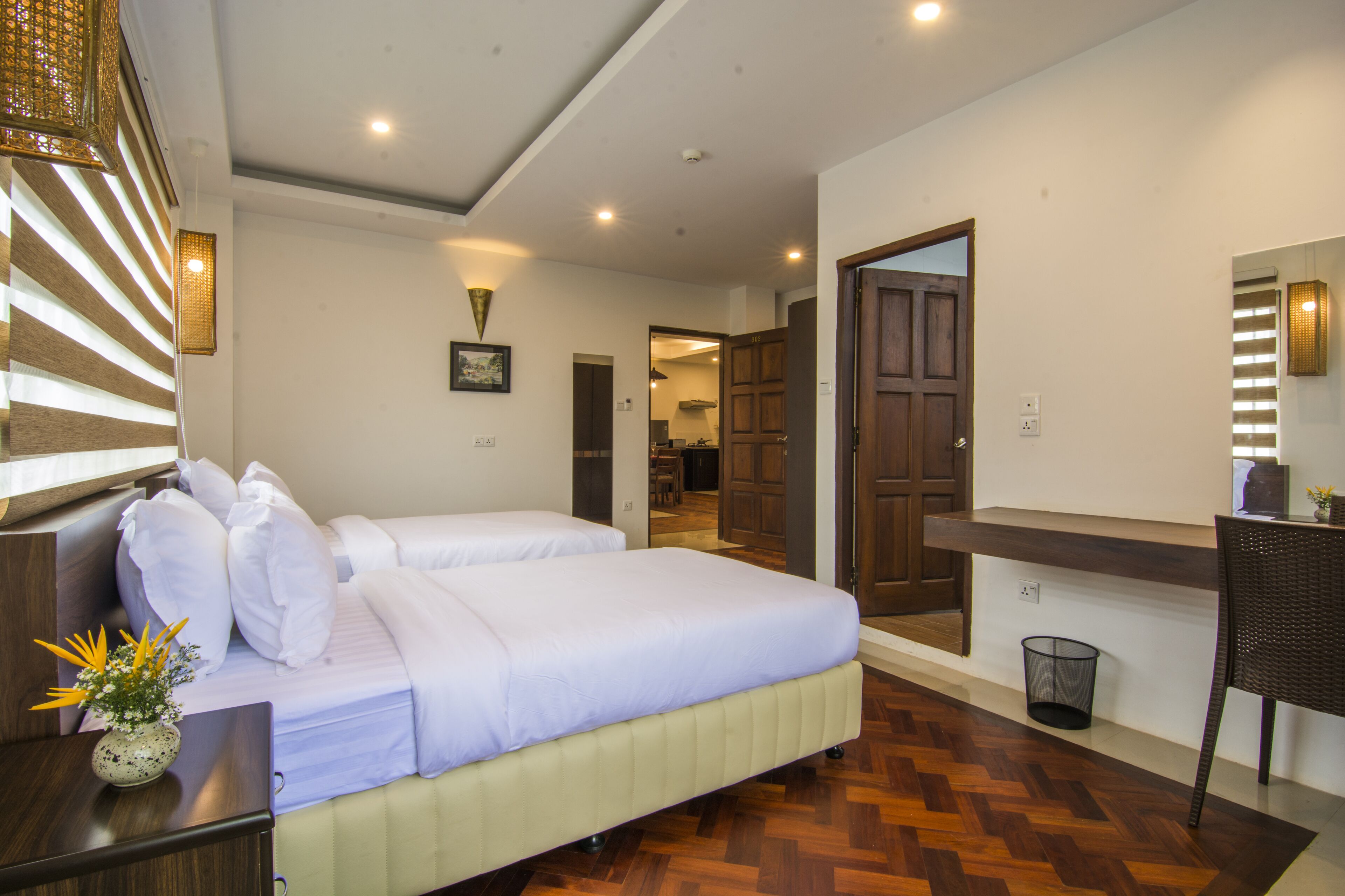 Habitación de hotel con dos camas, piso de madera, un escritorio con silla, un espejo y una puerta que da a otra habitación.
