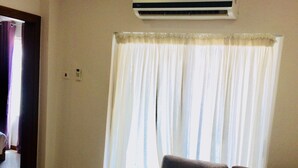 Una sala de estar con un sofá, un equipo de aire acondicionado, una ventana con cortinas y un piso de baldosas.