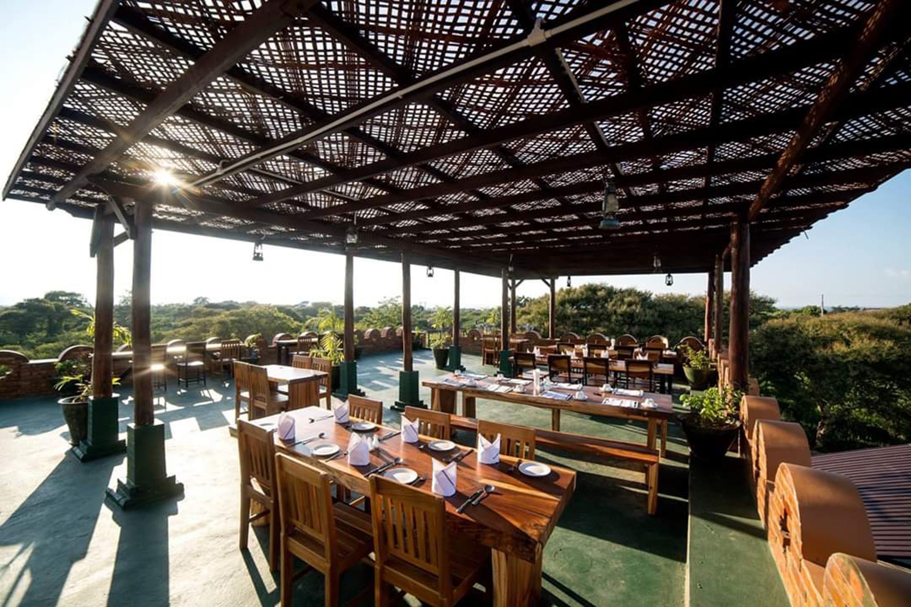 An outdoor dining area with wooden tables and chairs, a pergola structure, and a view of the surrounding landscape.