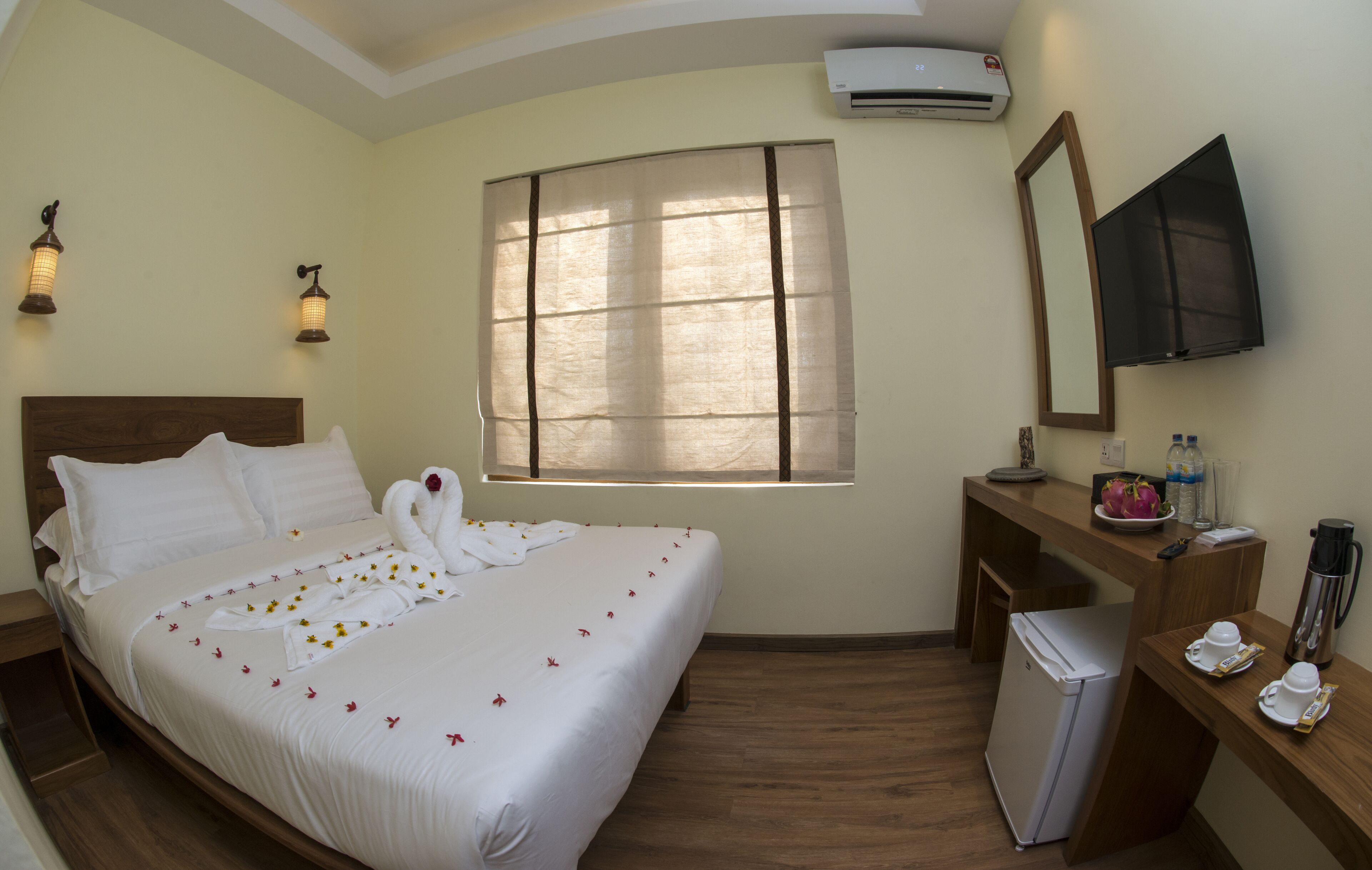 Habitación de hotel con cama, escritorio, refrigerador, espejo y ventana con persianas.