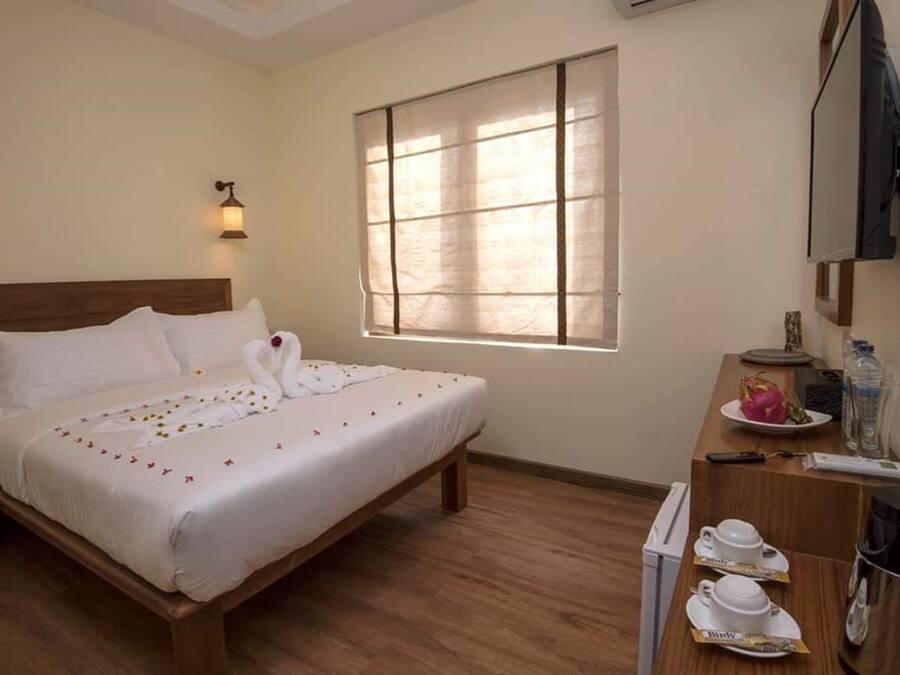 A neatly made bed with a white comforter and pillows, a wooden headboard, a large window with blinds, a bedside table with a lamp, a coffee maker, and a small TV on the wall.