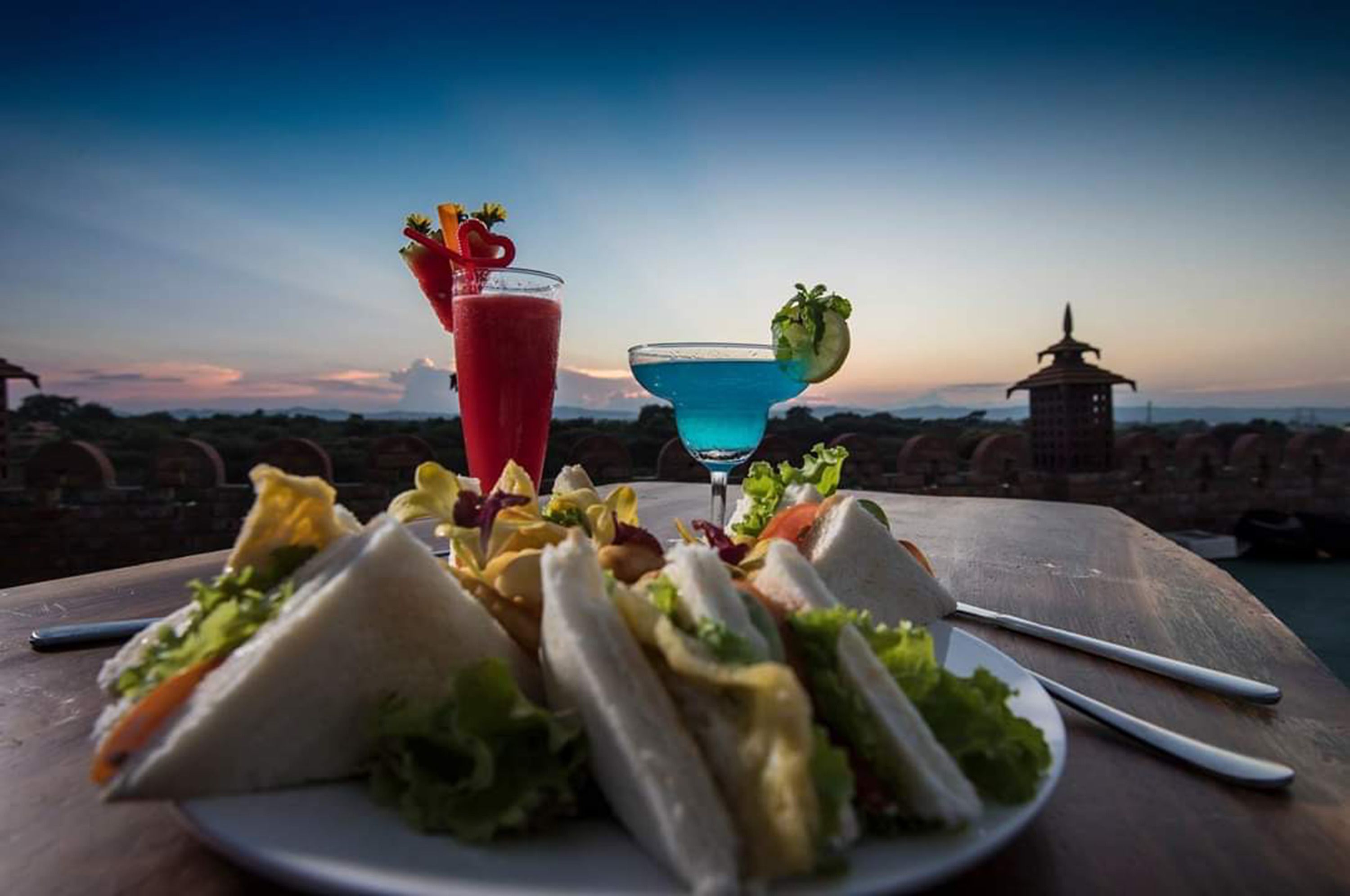 Een tafel met belegde broodjes, een rood drankje en een blauwe cocktail, voor een schilderachtige zonsondergang.