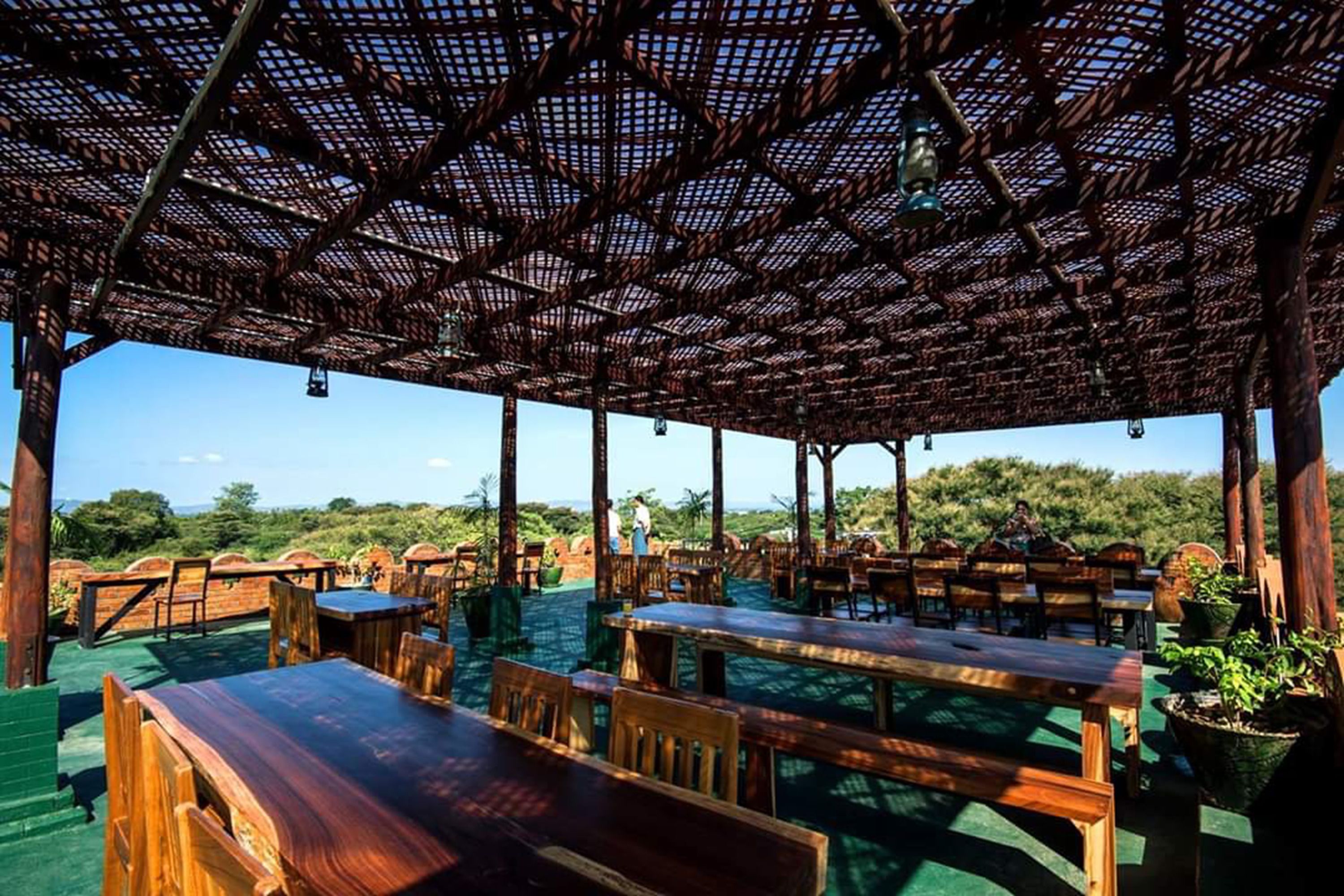 Un área de comedor al aire libre con mesas y sillas de madera, una estructura de techo tejido y vistas del paisaje circundante.