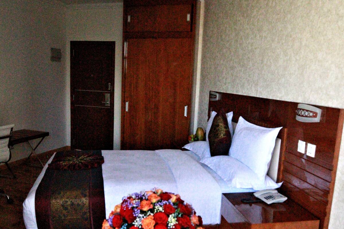 Ein Hotelzimmer mit Bett, Holz-Kopfteil, Nachttisch und einem kleinen Hocker mit Blumenarrangement.