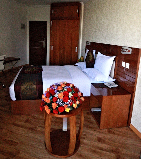 Quarto de hotel com cama, cabeceira de madeira, mesa de cabeceira e um pequeno banco com arranjo floral.