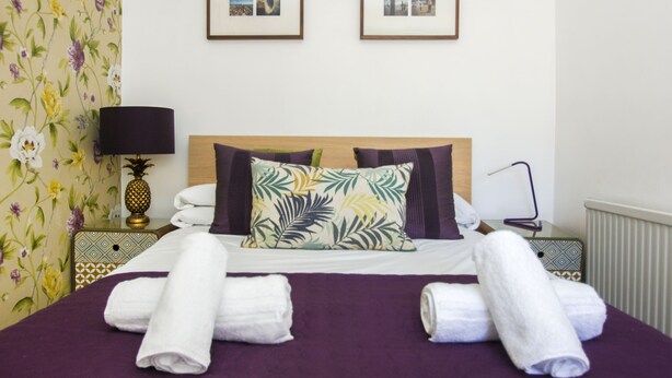 Un lit bien fait, recouvert d’une couvre-lit violette, deux tableaux encadrés accrochés au mur et un motif floral sur le papier peint d’un des murs.