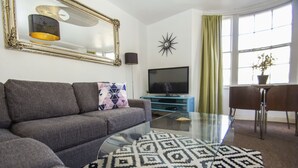 Un salon avec un canapé gris, une table basse en verre, un téléviseur à écran plat, un miroir décoratif et une plante en pot près de la fenêtre.