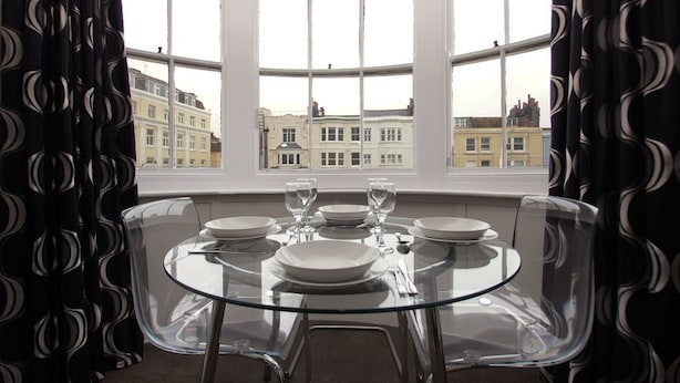 A dining area with a round glass table set for four, large windows with a view of buildings, and black curtains with a circular pattern. - New Steine Apartment Sea View by Brighton Holiday Lets (Brighton)