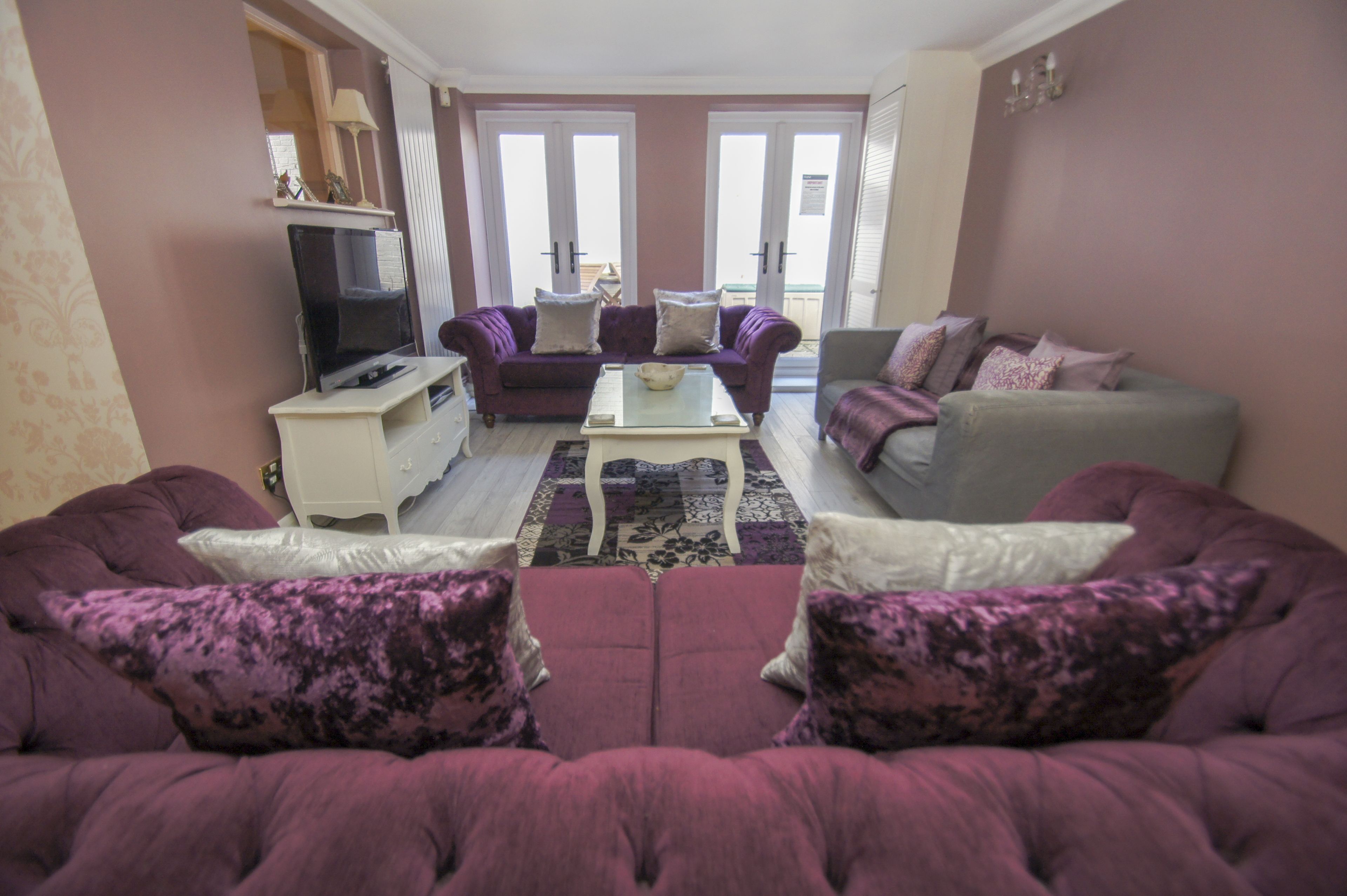 Ein Wohnzimmer mit zwei lilafarbenen Sofas, einem weißen Couchtisch und einem grauen Sofa. Ein Fernseher steht auf einer weißen Konsole und ein gemustertes Teppich liegt auf dem Boden.