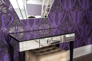 A bathroom vanity with a mirror, a beige cushioned stool, and a purple patterned wall. - Chocolate Box Central by Brighton Holiday Lets (Brighton)