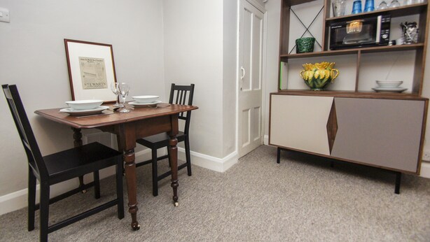 A dining area with a wooden table, black chairs, a framed certificate on the wall, and a cabinet with glassware and a vase of flowers. - Chocolate Box Central by Brighton Holiday Lets (Brighton)