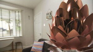 Sala de estar com design moderno, destacando uma grande escultura de planta metálica, um sofá escuro e almofadas coloridas com estampas.