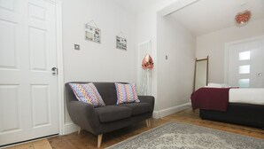 Quarto com sofá cinzento, almofadas estampadas, tapete com padrão, radiador, espelho e cama com manta vermelha.