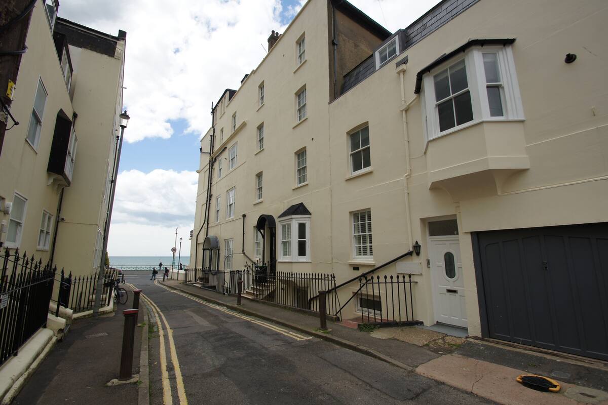 A narrow street with buildings on both sides, a visible entrance to a property, and a clear view of the sea in the distance.
