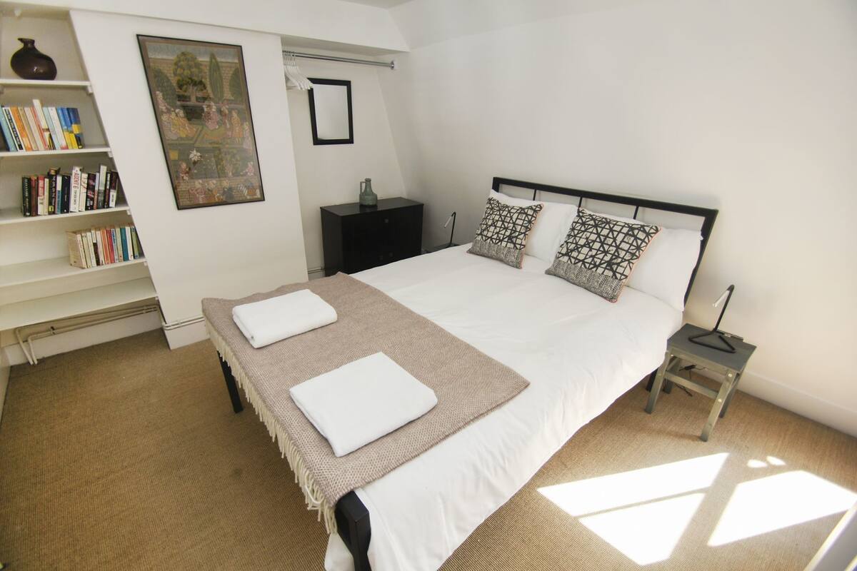 A neatly arranged bedroom with a large bed, a bedside table, a nightstand, a bookshelf, and a framed picture on the wall.