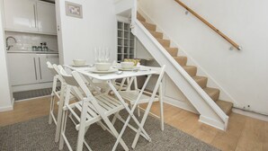 A compact kitchen with white cabinetry, a dining area with white chairs and a table set for four, and a wooden staircase.