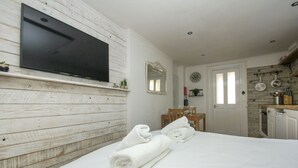 A hotel room with a wooden wall, a flat-screen TV mounted above a bed, a small table with a chair, and a kitchen area with a stove and utensils. - Brunswick Garden Studio Patio by Brighton Holiday Lets (Hove)