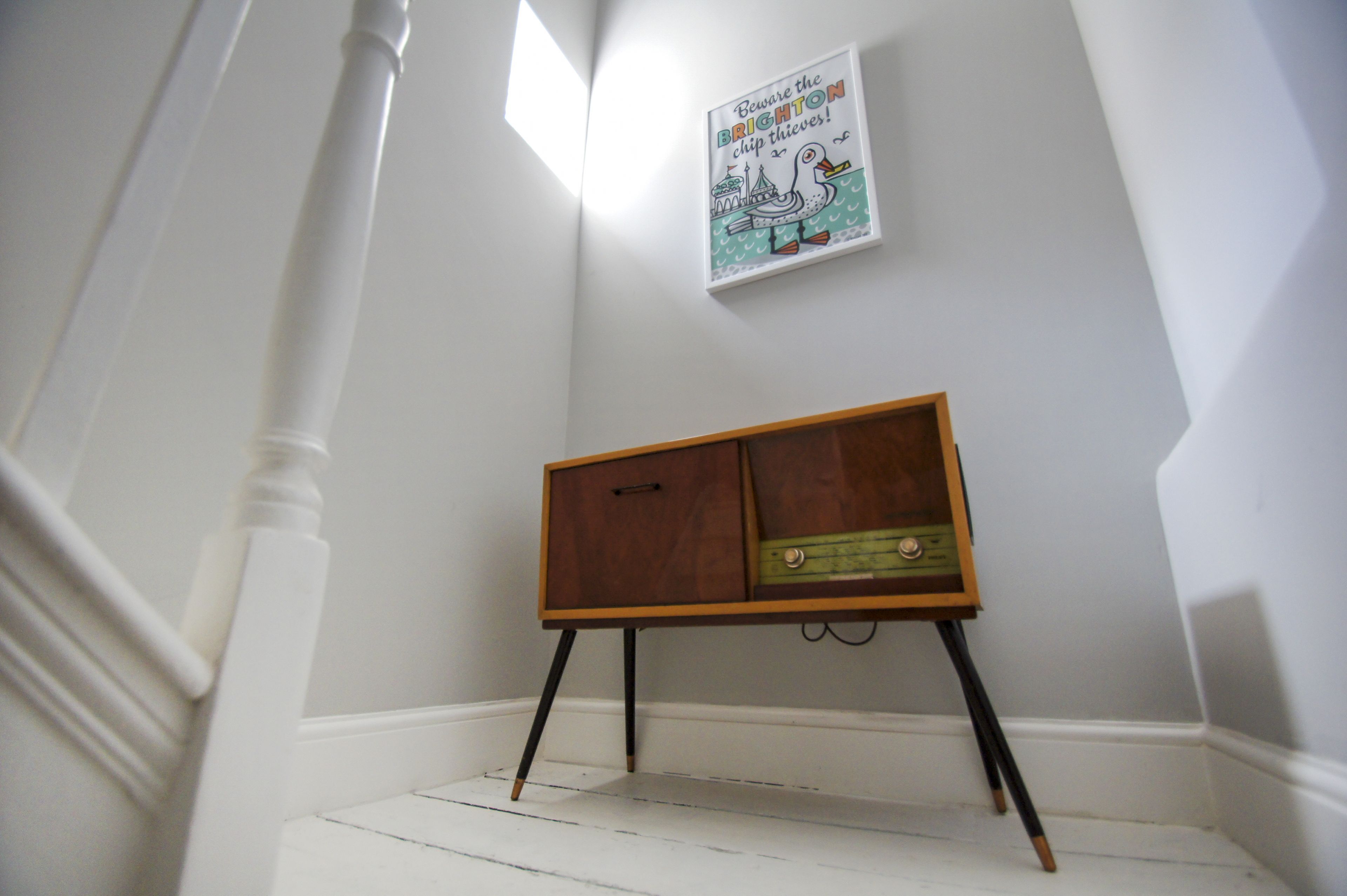 A corner with a white staircase, a wall-mounted poster, and a vintage wooden cabinet with a drawer.