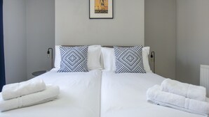 Een tweepersoonsbed met witte lakens en gekleurde kussens, een ingelijst kunstwerk aan de muur, en opgerolde handdoeken op het bed.