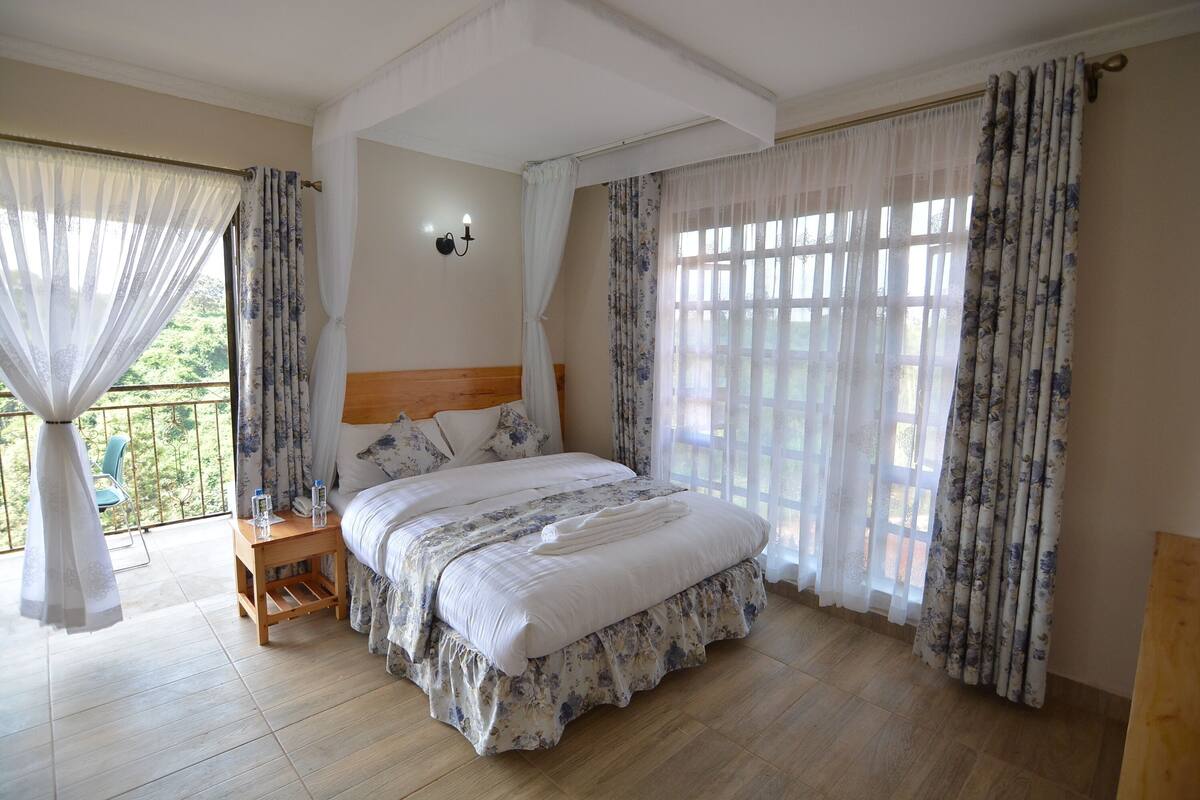 Quarto com cama grande, mesa de cabeceira de madeira, luminária na parede e vista para a varanda.
