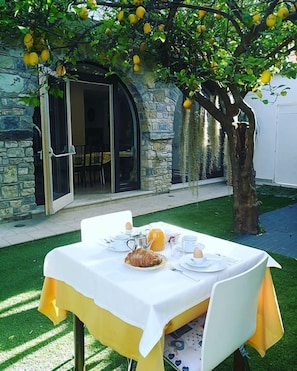 Un espacio de comedor al aire libre con una mesa preparada para el desayuno, un edificio de piedra al fondo y un limonero.
