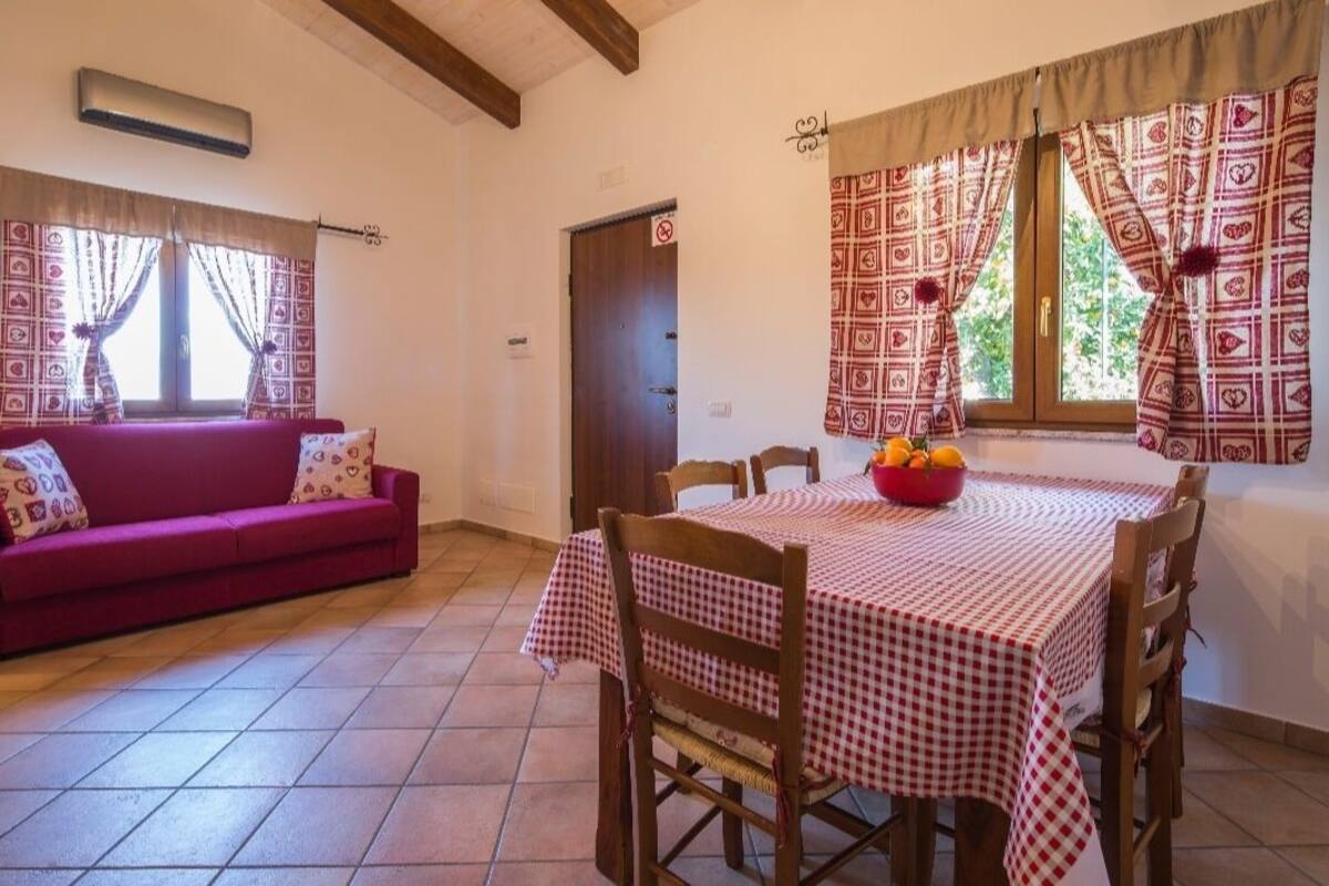 Un comedor con mantel a cuadros, un sofá rojo con cojines estampados y una ventana con cortinas decoradas.