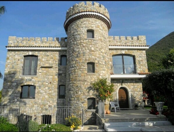 Un edificio parecido a un castillo de piedra, con una torre central, ventanas amplias y una entrada con portón.