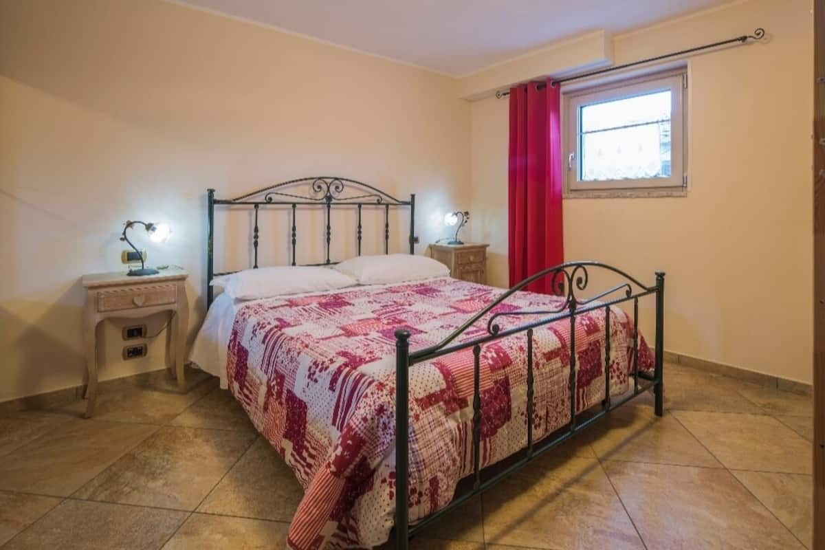 Una cama individual con estructura metálica, dos mesitas de noche con lámparas, una ventana con cortina roja y piso de baldosas.