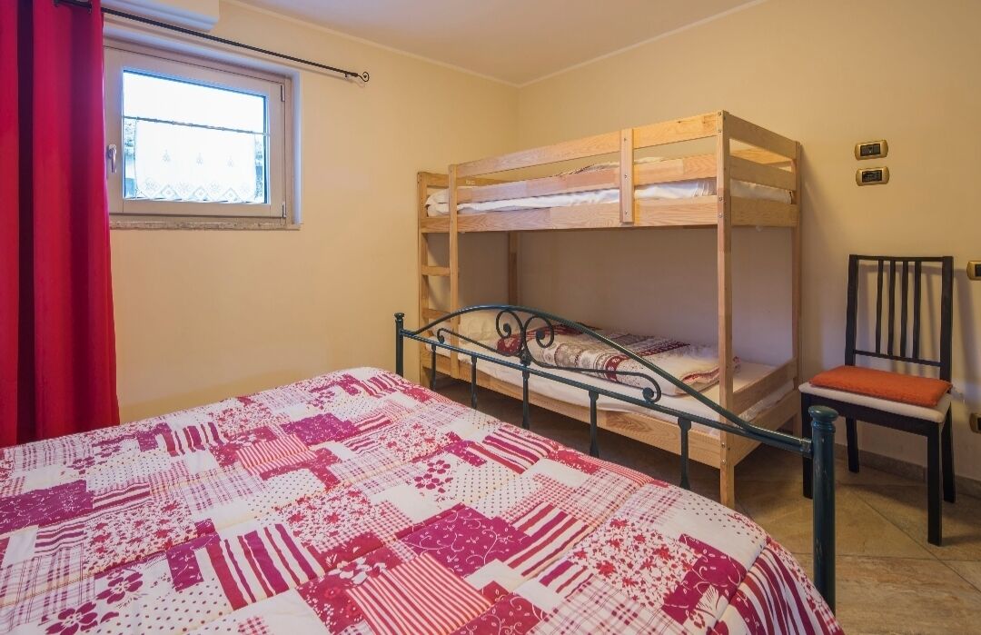 A bunk bed room with a quilted bedspread, a window with a view, and a chair.