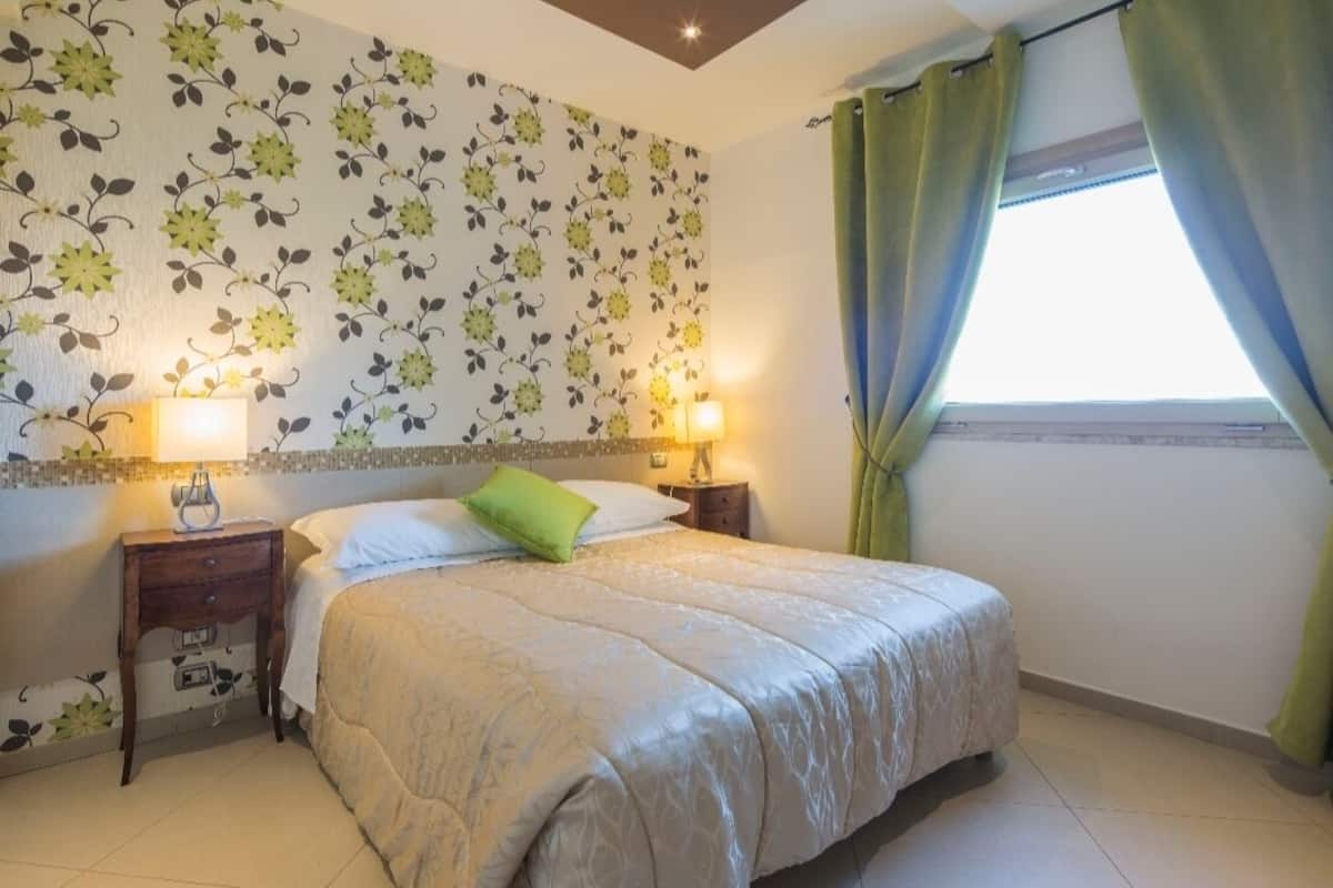 Un dormitorio con cama, dos mesitas de noche con lámparas, un papel tapiz floral y una ventana con cortinas.