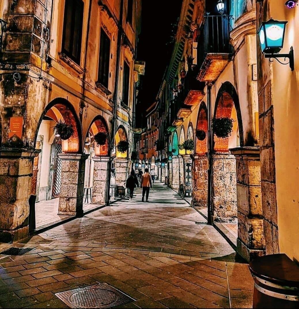 Una calle estrecha con edificios de piedra, puertas en arco y macetas colgantes con flores.