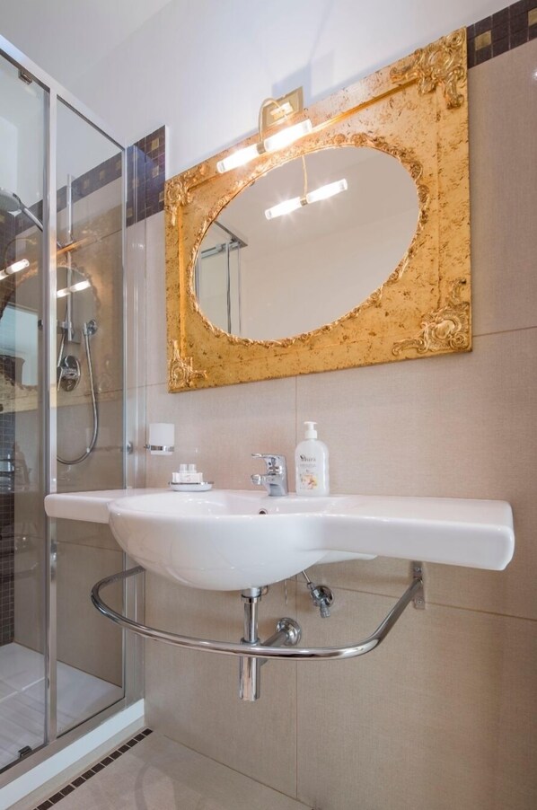 Une salle de bain avec un lavabo blanc, un miroir encadré de doré et une cabine de douche en verre.